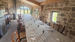 Una larga mesa de comedor dispuesta con platos, vasos y cubiertos en una sala rústica de paredes de piedra y vigas de madera, con vistas a la vegetación exterior.