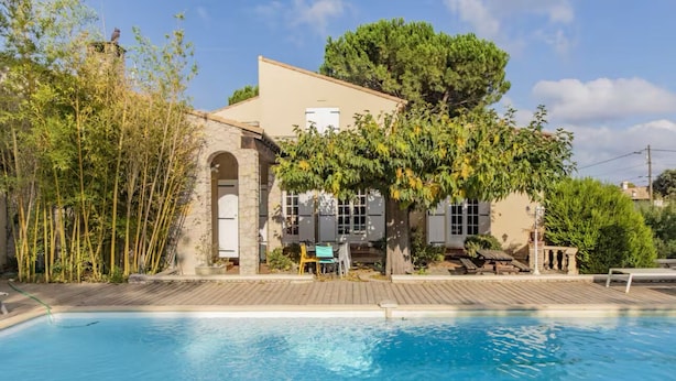 A poolside view of a building with a stone facade, a large tree, and a clear blue sky. - Bastide in Nîmes ⭐️⭐️⭐️ (Nîmes)