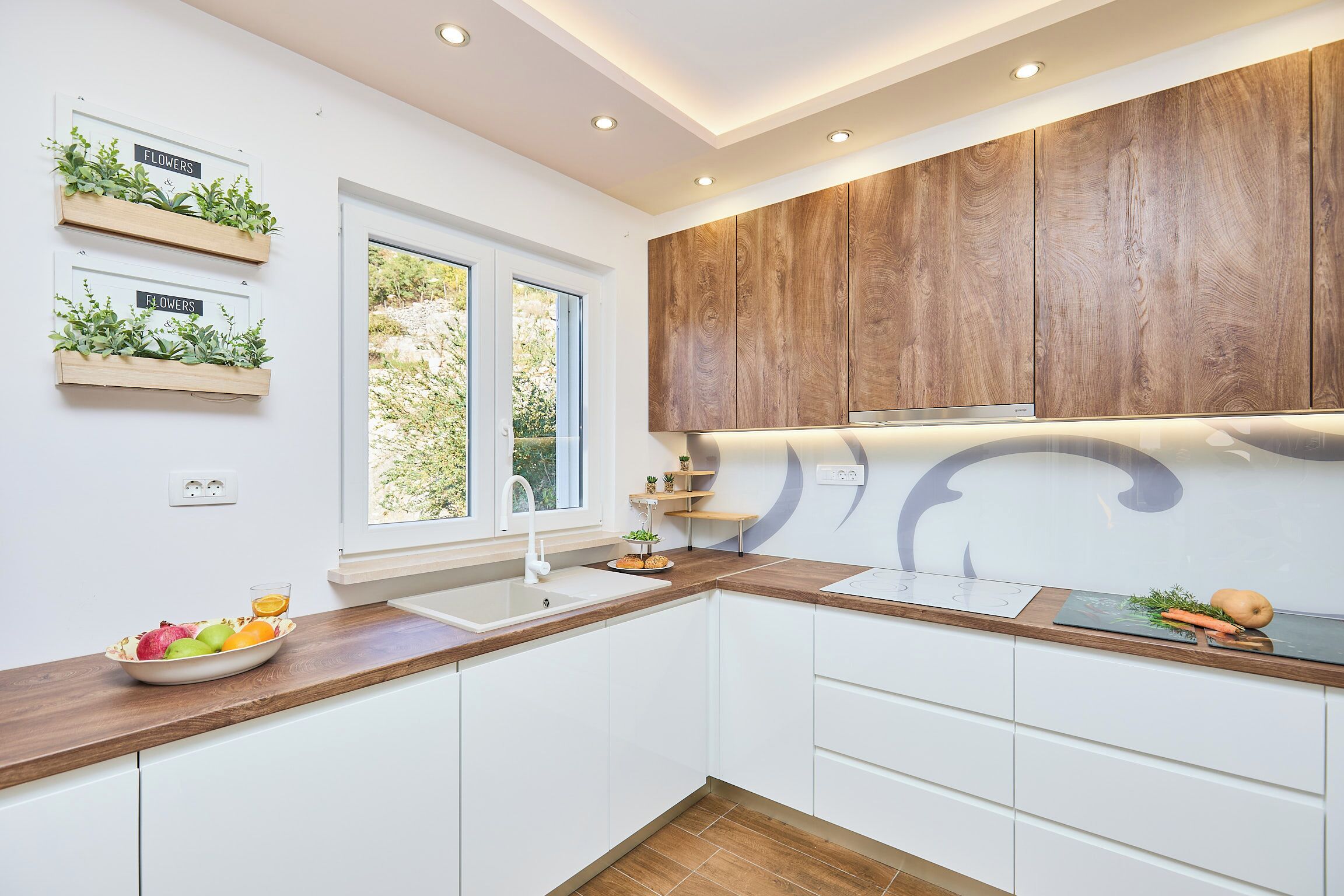 A modern kitchen with white cabinets, wooden countertops, and built-in wooden cabinets above the counter. There are two windows providing natural light, and a bowl of fruit on the counter.