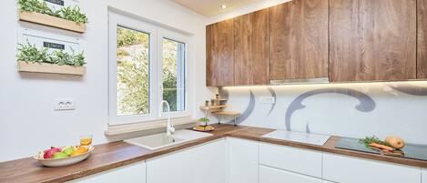 A modern kitchen with white cabinets, wooden countertops, and built-in wooden cabinets above the counter. There are two windows providing natural light, and a bowl of fruit on the counter.