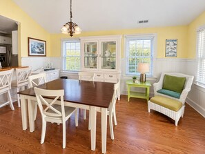 Un coin repas avec une table en bois et des chaises blanches, une petite table d’appoint avec une lampe, et un fauteuil en osier avec un coussin.