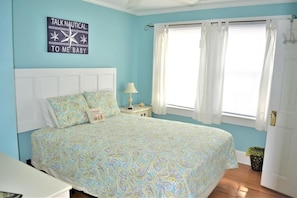 A bedroom with a large bed, a nightstand, a lamp, a window with curtains, and a wall sign. - Pink Pearl-Super cute pet friendly cottage, 3 bedroom 2 bath with a rocking chair front porch and plenty of parking (Kure Beach)