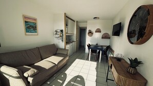 Een woonkamer met een bank, een eettafel met stoelen, een keukenhoek en een aan de muur bevestigde televisie.