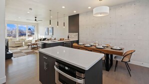 A modern kitchen with a large island, dining area with wooden table and chairs, and a view of the outdoors through large windows. - Mountain View Home w/ King Beds & Rooftop Deck (Fort Collins)