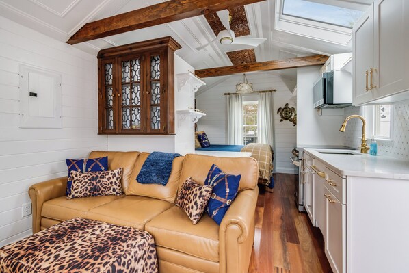 A spacious living room with a tan leather sofa, patterned cushions, and a wooden ceiling beam. A kitchen with white cabinets and a gold faucet is visible in the background. - Charming Tiny Home in Rehoboth Beach with a Water View (Rehoboth Beach)