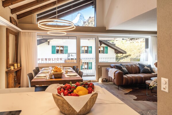 A dining area with a table set for four, a fruit bowl, and a view of a mountain through large windows.
