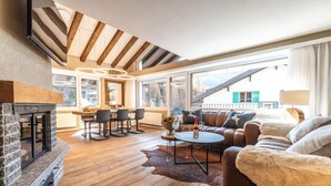 Ein modernes Wohnzimmer mit Kamin aus Naturstein, Holzbalken, einer Ledersofa, einem runden Couchtisch und einem Essbereich mit Bergblick.