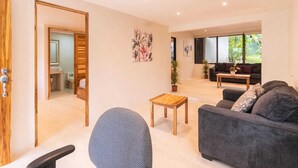 A modern living room with a sofa, a coffee table, and a chair. There is a door leading to a bathroom, and a window with a view of greenery outside. - Surfside Apartments Mal Pais (Cóbano)