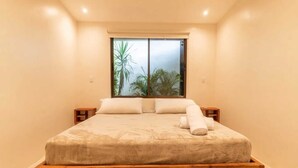 A bedroom with a wooden bed, two bedside tables, a window with a view of palm trees, and a ceiling light. - Surfside Apartments Mal Pais (Cóbano)