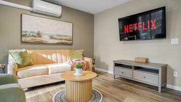 A living room with a beige sofa, a round wooden coffee table, a flat-screen TV, and a wall-mounted air conditioner.