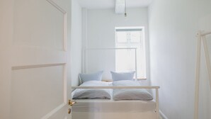 A narrow, white room with a bed at the end, a window with blinds, and a door with a brass handle.