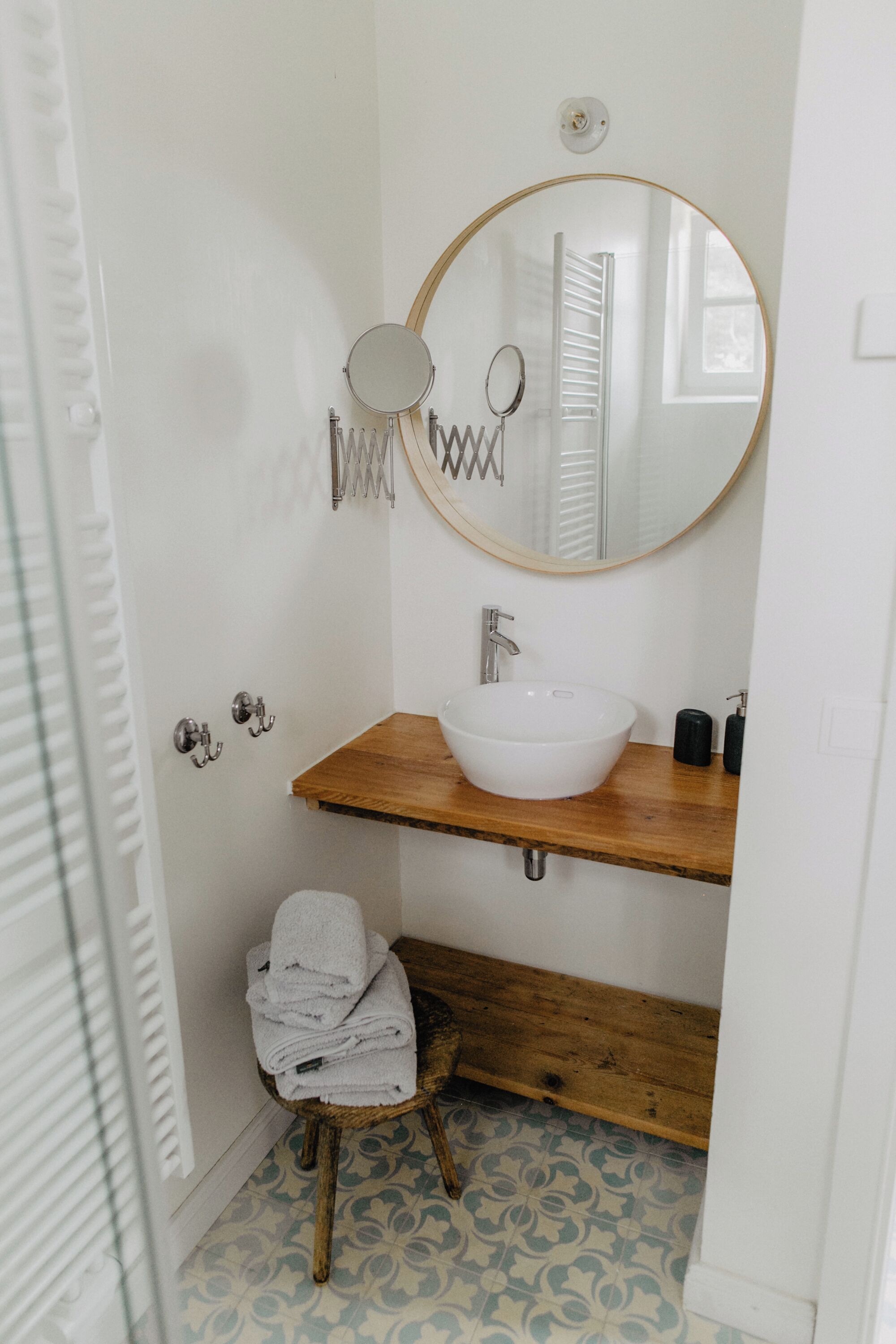 A bathroom with a round mirror, a wooden shelf, a white sink, and a small stool with towels.