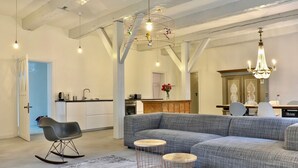 A spacious living area with a grey sectional sofa, a rocking chair, and a dining table with chairs. There are two small round tables with wire frames and a chandelier hanging from the ceiling.