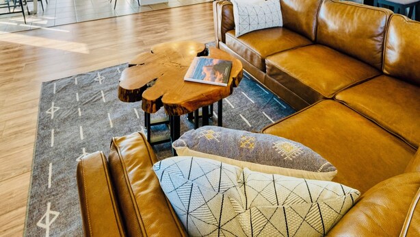 Een ruime woonkamer met een bruine leren bank, een houten salontafel, en een eethoek met een tafel en stoelen.