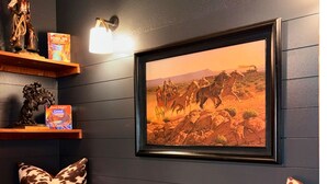Een kamer met een donkere muur, een lijst met een schilderij van een woestijnlandschap, een houten plank met decoratieve objecten en een comfortabele bank met een kussen met een patroon.