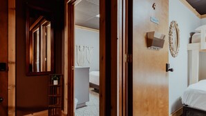 A room with a bed, a wooden door, a shelf with a plant, and a wall-mounted mirror.