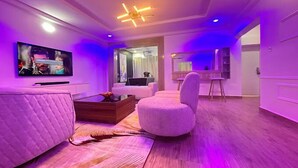 A modern living room with a flat-screen TV, a sofa, a coffee table, and a round ottoman. - DE CALMA APARTMENTS (Abuja)