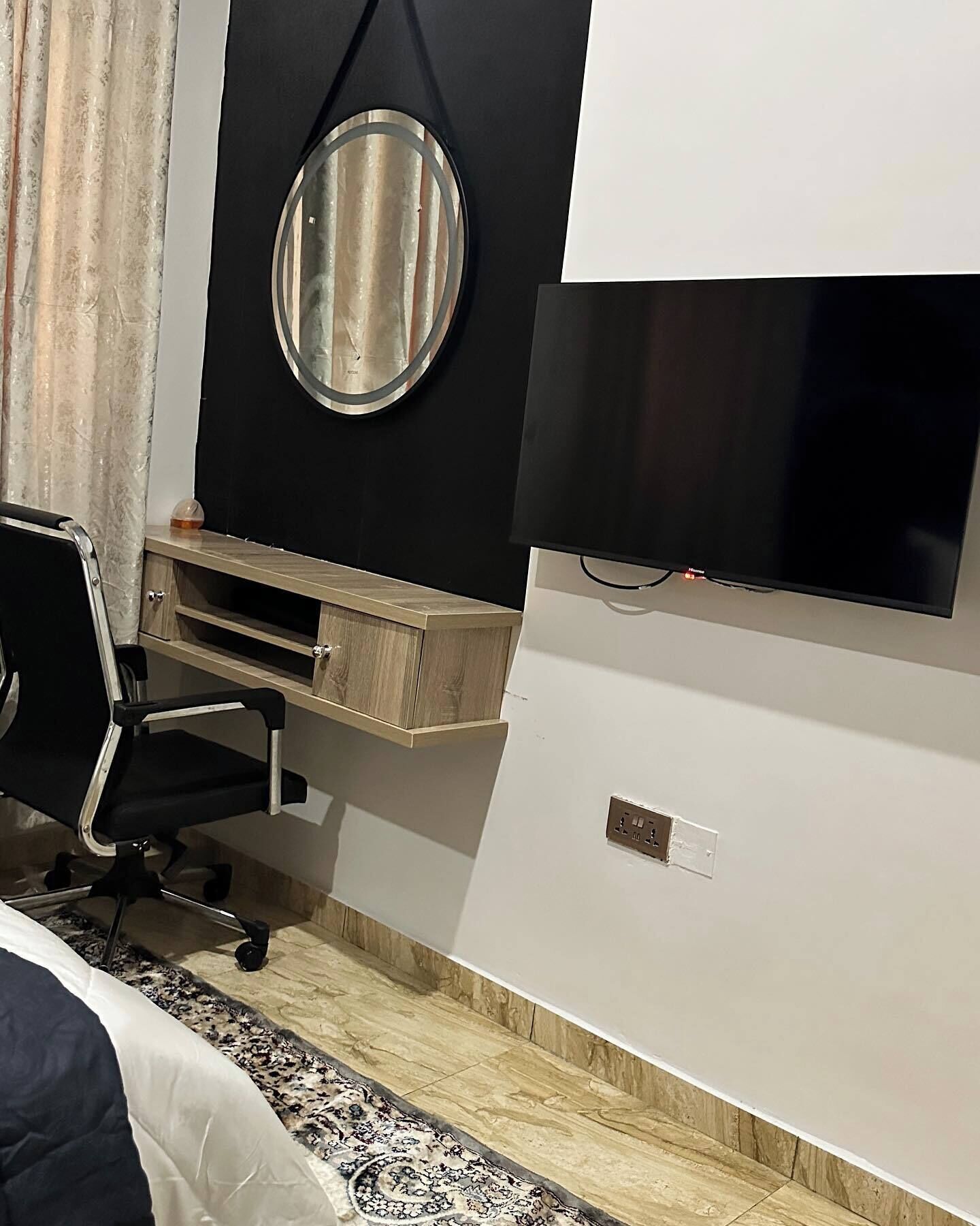 A modern bedroom with a bed, a desk, a chair, a TV, and a mirror.