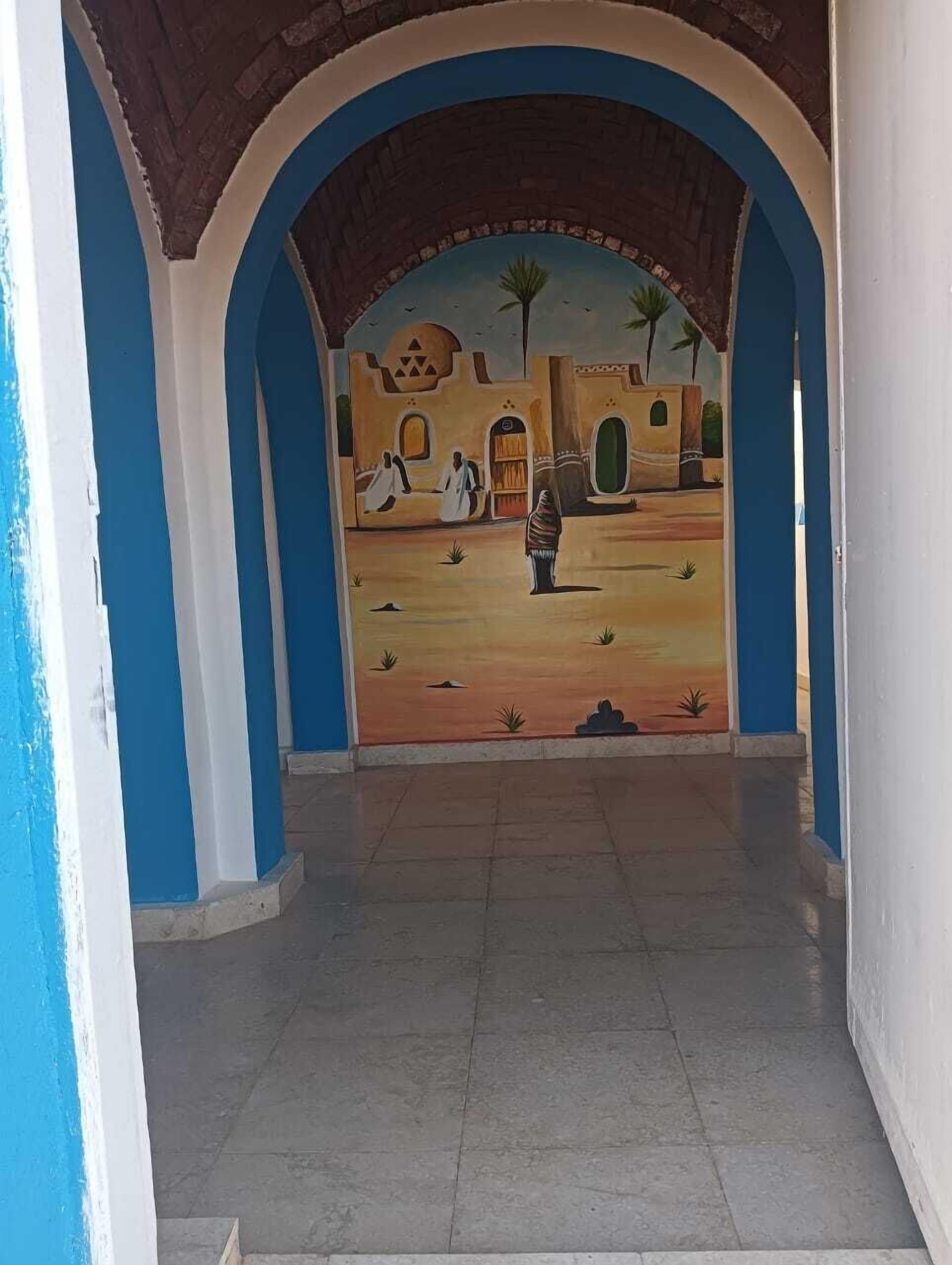 Een hal met blauw-witte wanden, een betegelde vloer en een muurschildering van een traditionele scène met mensen en palmbomen.