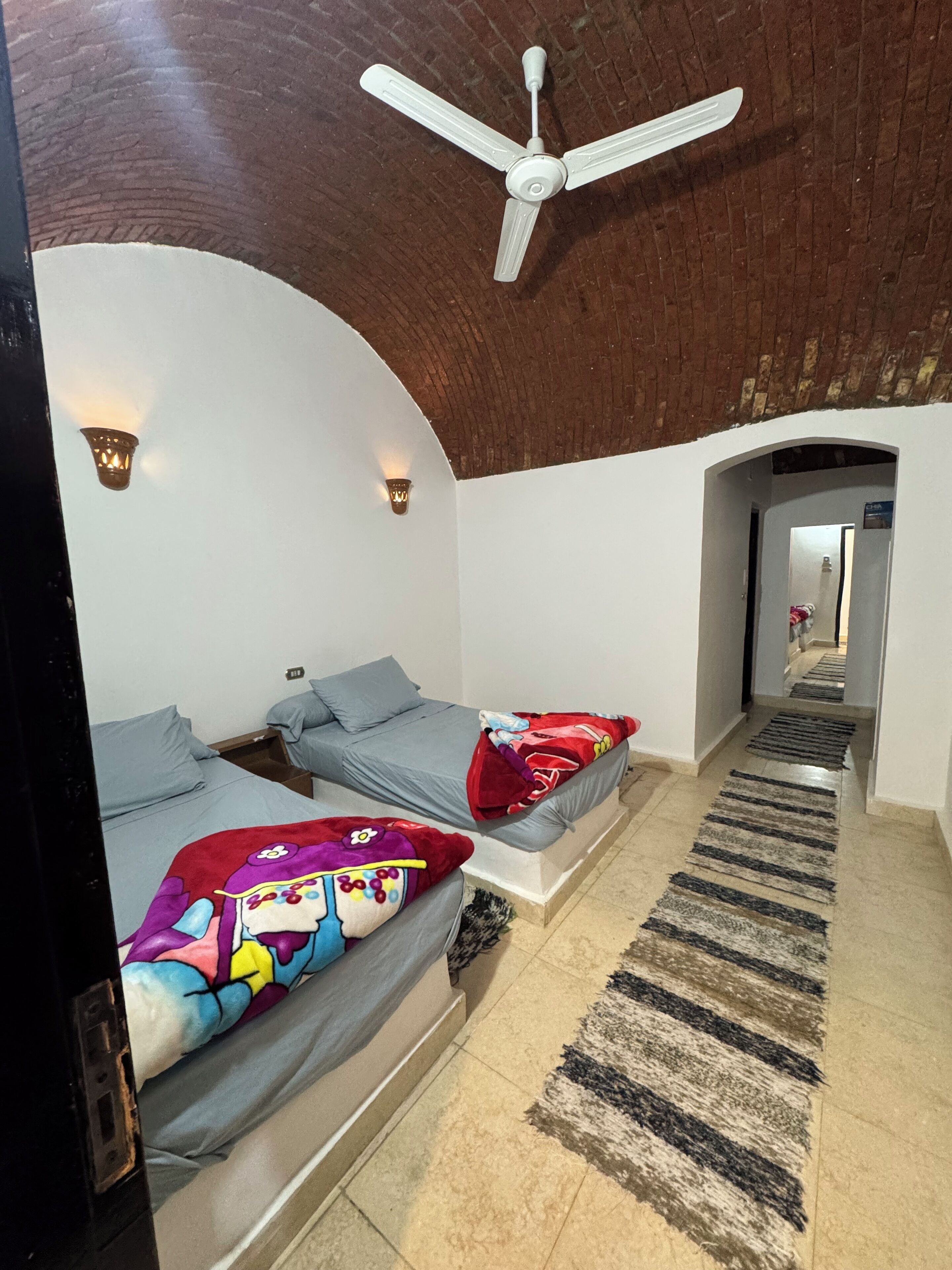 A room with two beds, a ceiling fan, and a hallway leading to another room.