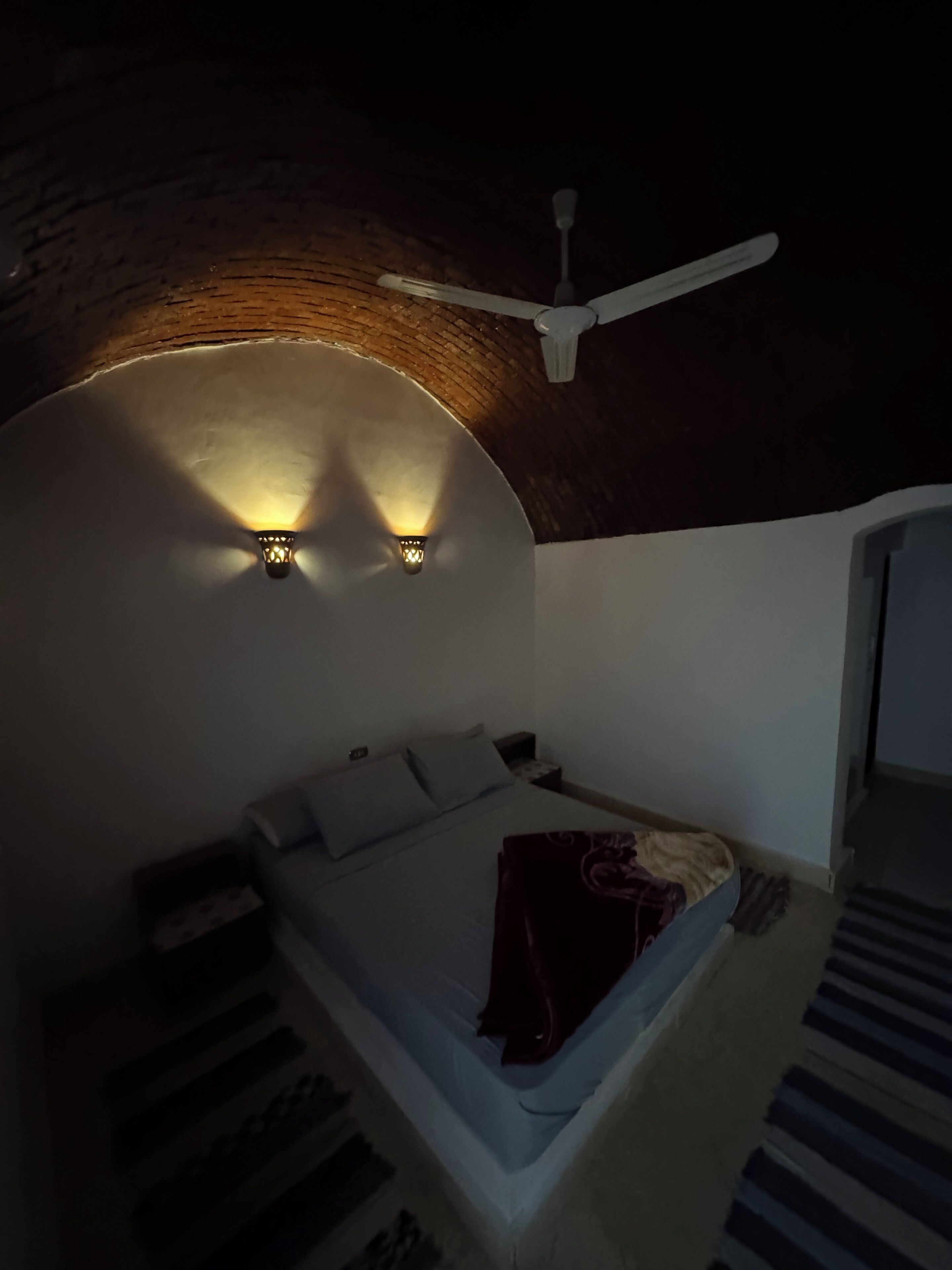 Una stanza scarsamente illuminata con un letto, una ventola a soffitto e due luci a parete.
