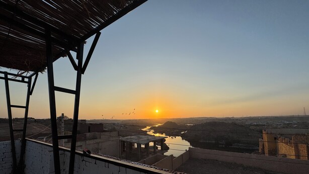 Blick auf einen Sonnenuntergang über einem Fluss von einem Balkon mit einer Backsteinmauer und einer Metallkonstruktion.