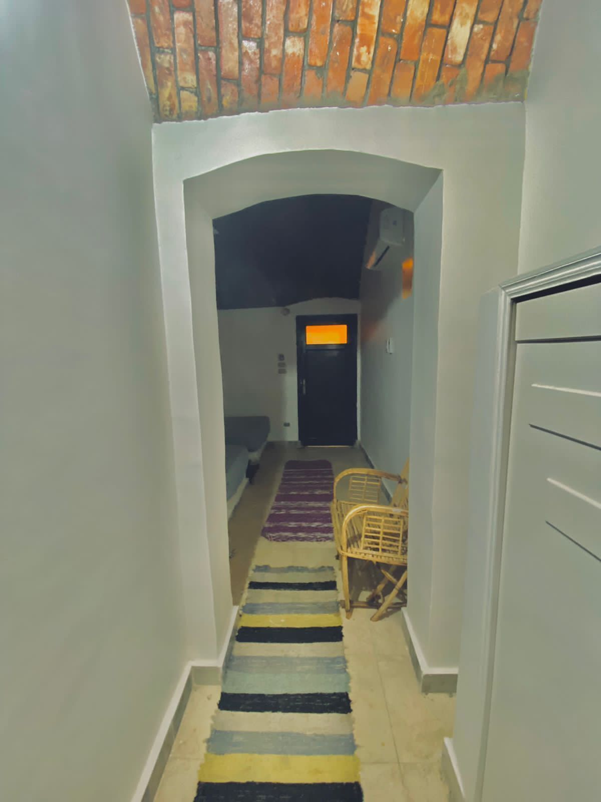 A narrow hallway with a striped rug, a wicker chair, and a door with an orange light.