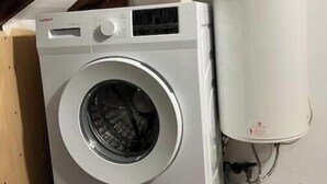 A compact, white washing machine in a small, enclosed space with a wooden ceiling and a white water heater mounted on the wall.