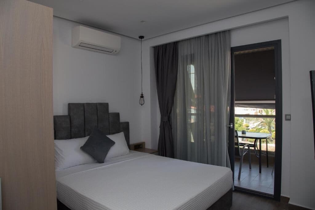 Quarto moderno com cama grande, cabeceira, mesa de cabeceira e vista para uma varanda com mesa e cadeiras.