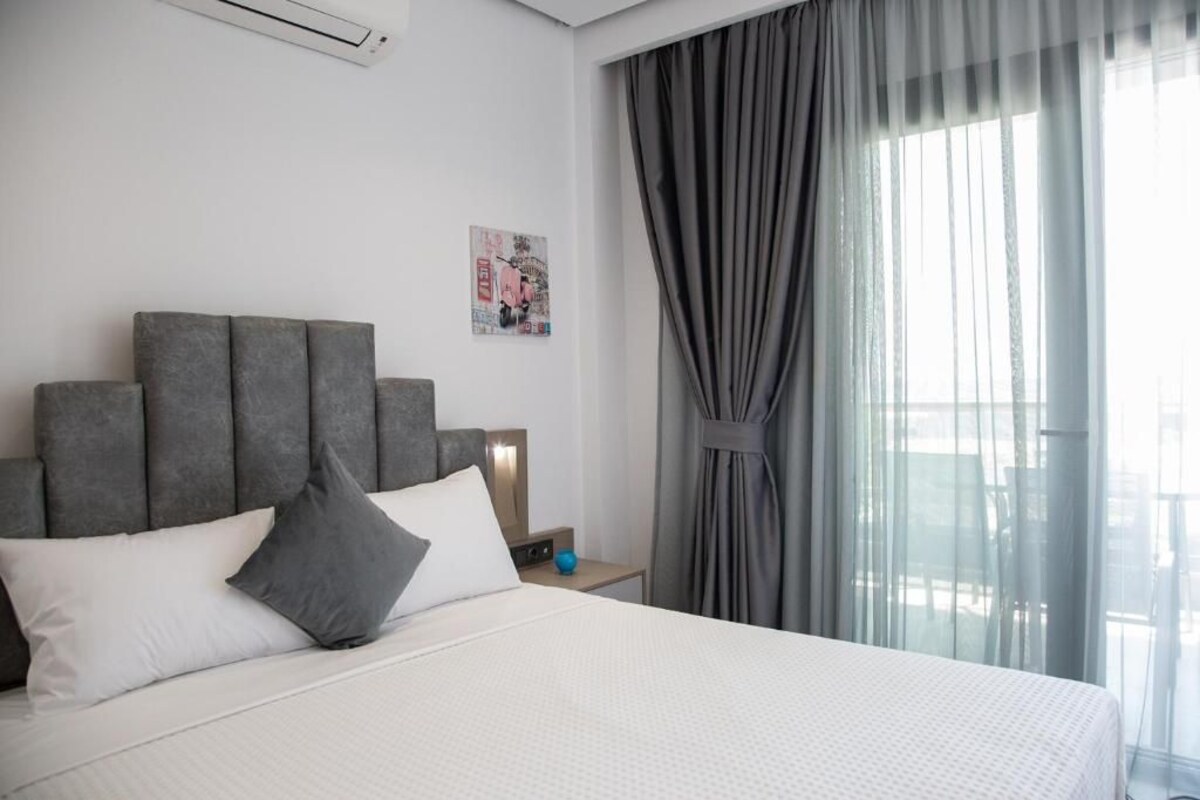 Une chambre d’hôtel moderne avec un grand lit, une tête de lit grise et une vue sur la ville à travers des rideaux transparents.