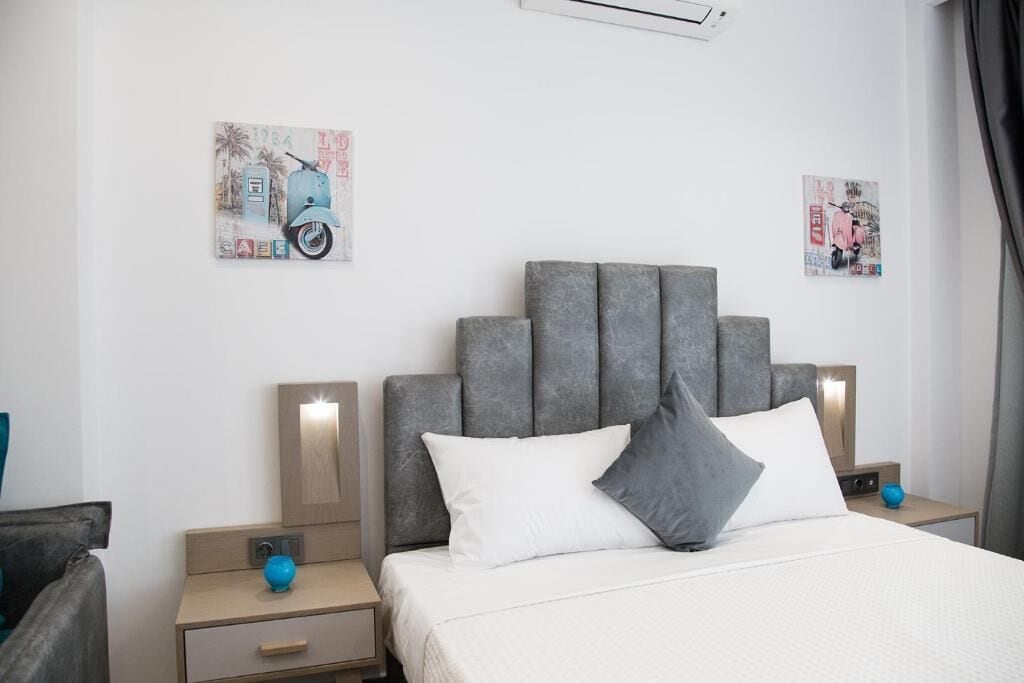 A modern hotel room with a grey upholstered headboard, a bed with white linens, two bedside tables with lamps, and artwork on the walls.