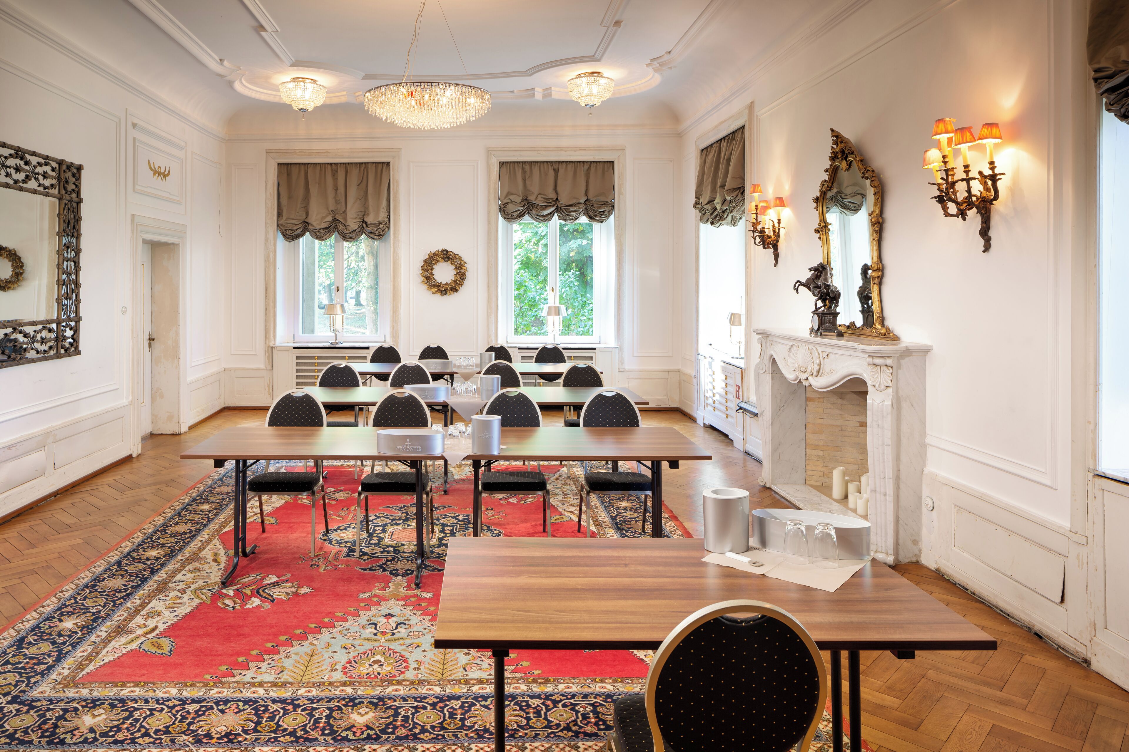 A formal dining room with a long table, multiple chairs, a patterned rug, and a fireplace with wall sconces.