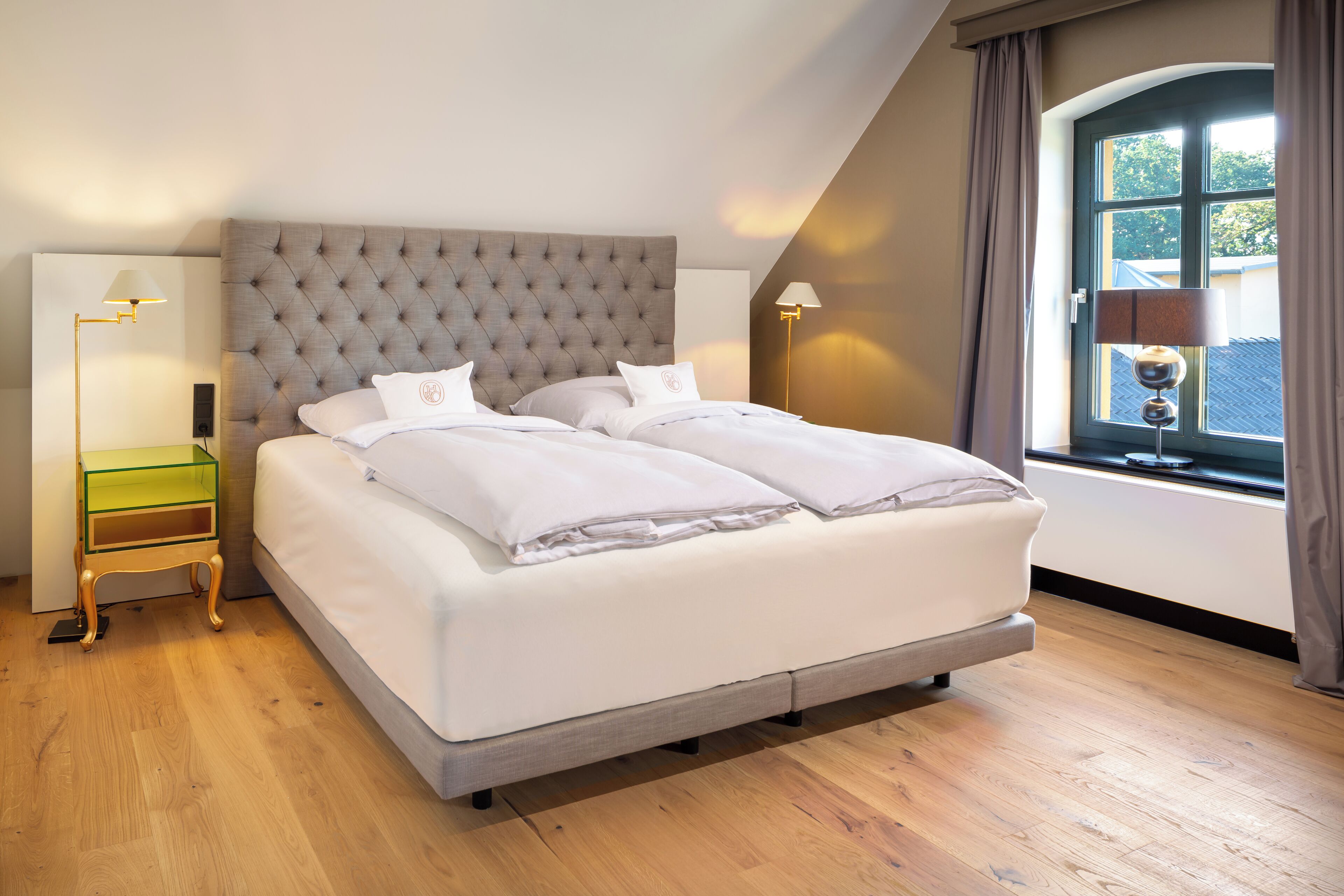 A neatly made bed with a tufted headboard, a bedside table with a lamp, and a window with curtains.