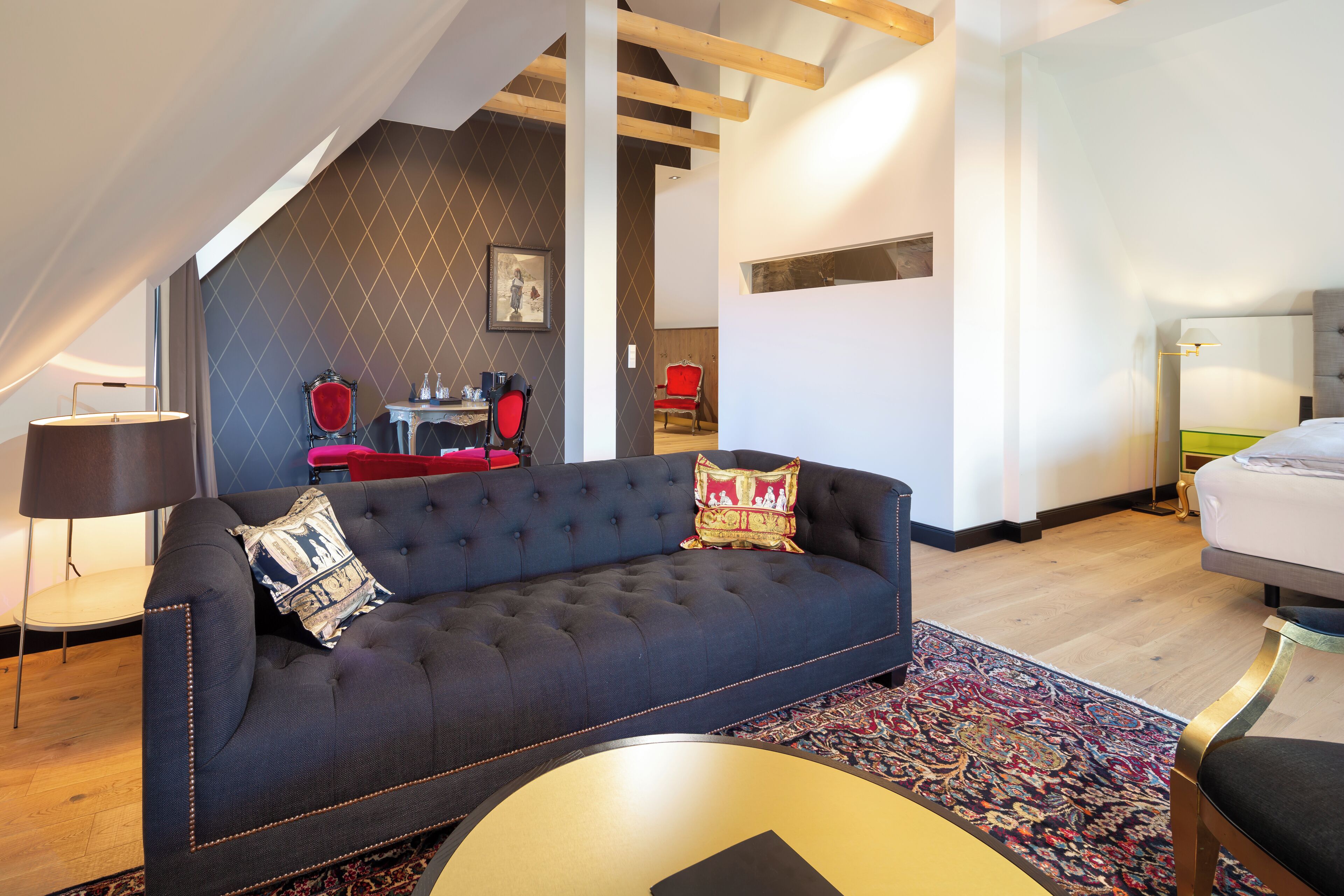 A modern living room with a dark blue sofa, a red chair, and a small table. There is a patterned rug on the floor and a lamp on a side table.
