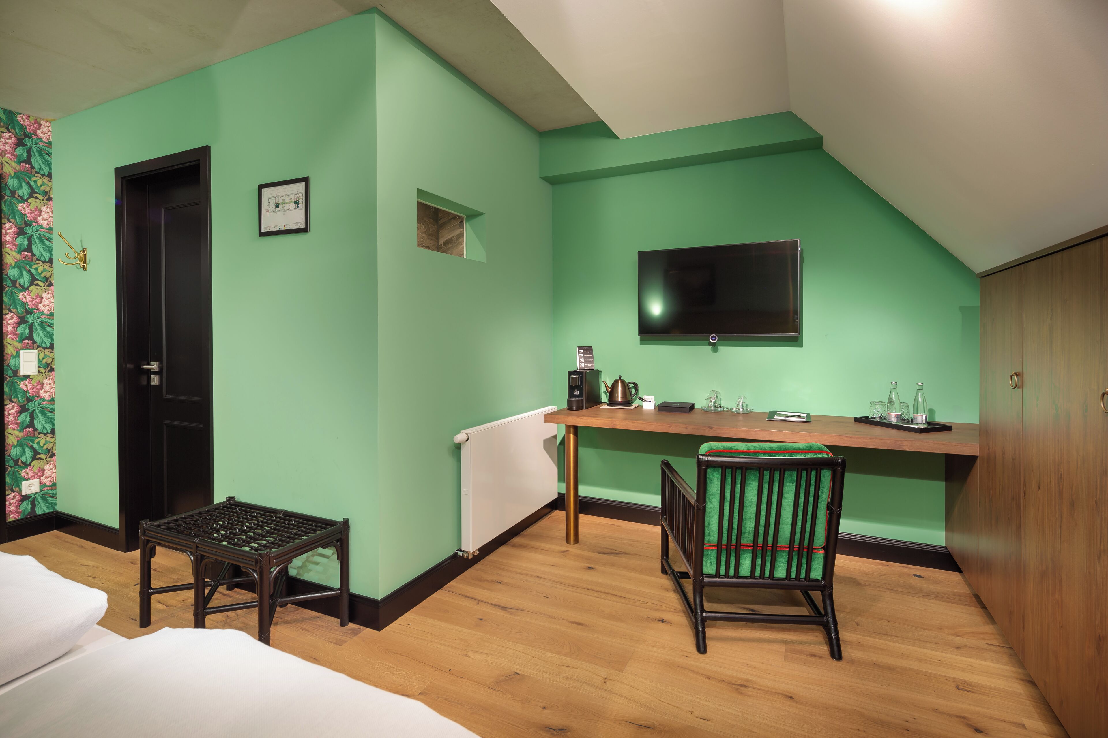 A room with a green wall, a wooden desk, a chair, a bed, and a television.
