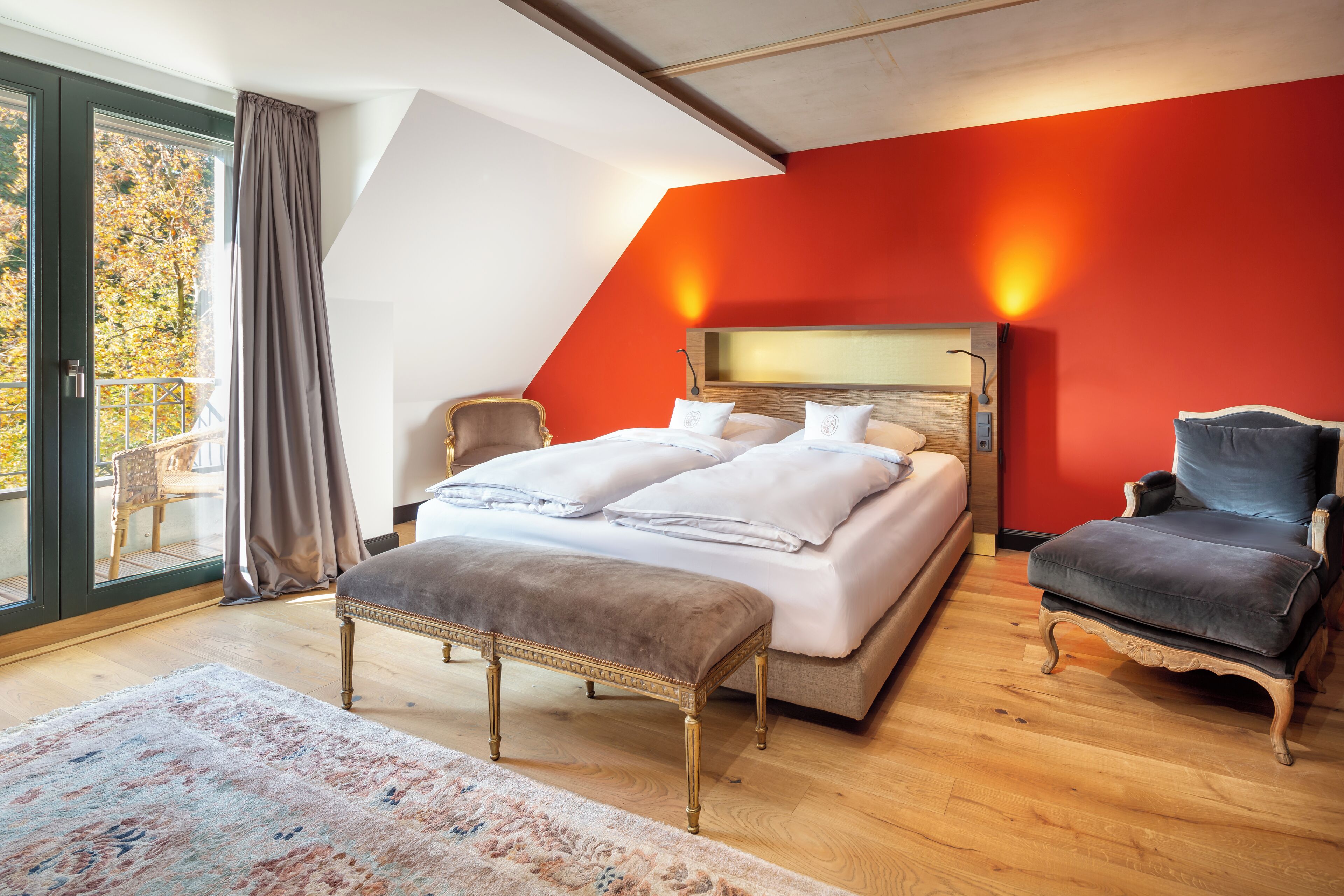 Un dormitorio con una cama, dos sillas, un banco, un ventanal grande y una pared roja.