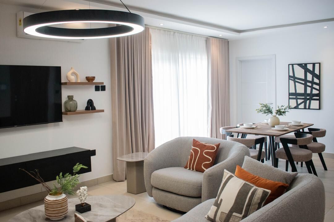 Een moderne woonkamer met een flatscreen televisie, grijze banken, een eettafel en een groot raam met gordijnen.