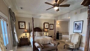 Ein Schlafzimmer mit einem Himmelbett, zwei Sesseln, einem Schreibtisch mit Lampe und einem Deckenventilator.