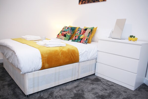Un lit bien fait, recouvert d’une couverture jaune et agrémenté d’oreillers blancs, une table de chevet blanche avec un ordinateur portable et une petite plante, et un sol recouvert d’un tapis gris.
