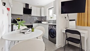 A compact kitchen with a washing machine, a dining table with chairs, a TV mounted on the wall, and a window with curtains. - South Harrow (Harrow)