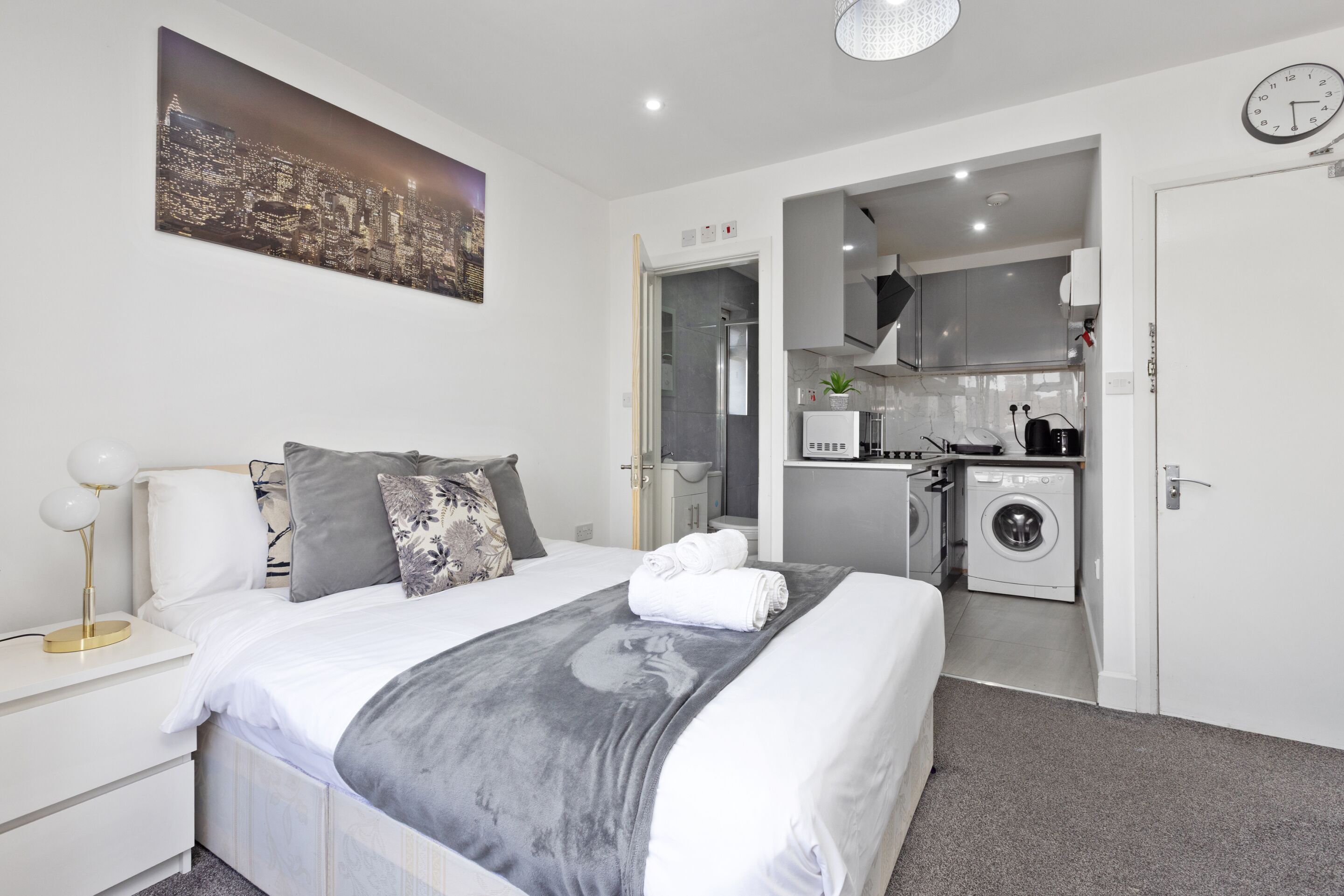 A modern bedroom with a large bed, a bedside table, a nightstand, a wall-mounted clock, and a cityscape painting.