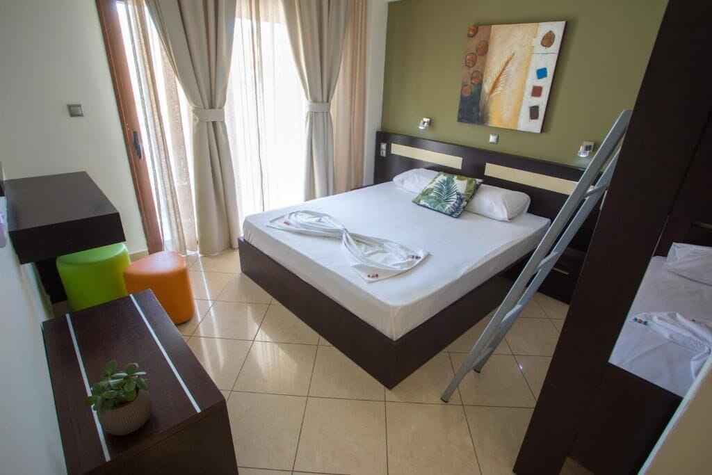 A hotel room with a bed, a bedside table with a plant, a window with curtains, and a painting on the wall.