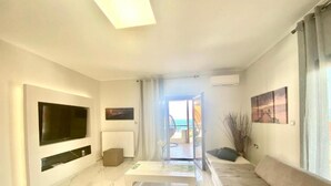 A modern living room with a large flat-screen TV, a sofa, a coffee table, and a view of the outdoors through a glass door. - Villa Amalia Sun Beach (Volvi)