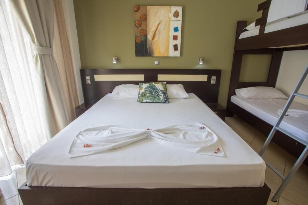 A hotel room with a single bed, a bunk bed, a painting on the wall, and a neatly made bed with towels.