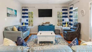 A living room with a grey sofa, a blue and white striped curtain, a white coffee table, and a black leather chair. - Book Coastal Cottage – Short Drive to the Beach! (Gulf Shores)