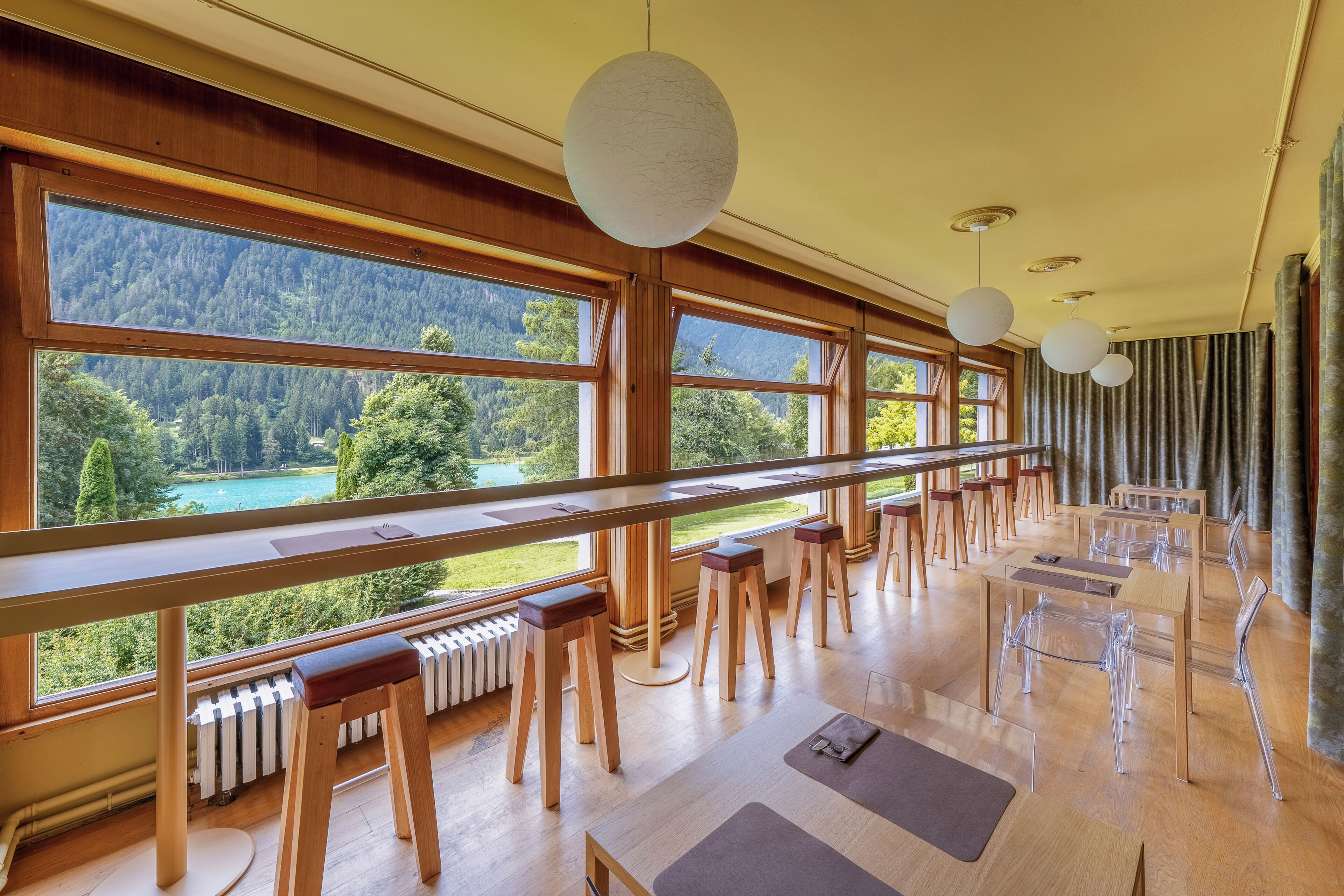 A spacious room with large windows offering a view of a forested area and a lake. There are wooden tables and chairs arranged for dining.