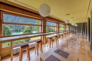 A spacious room with large windows offering a view of a forested area and a lake. There are wooden tables and chairs arranged for dining. - Hotel Auronzo Dolomites, Affiliated by Meliá (Auronzo di Cadore)