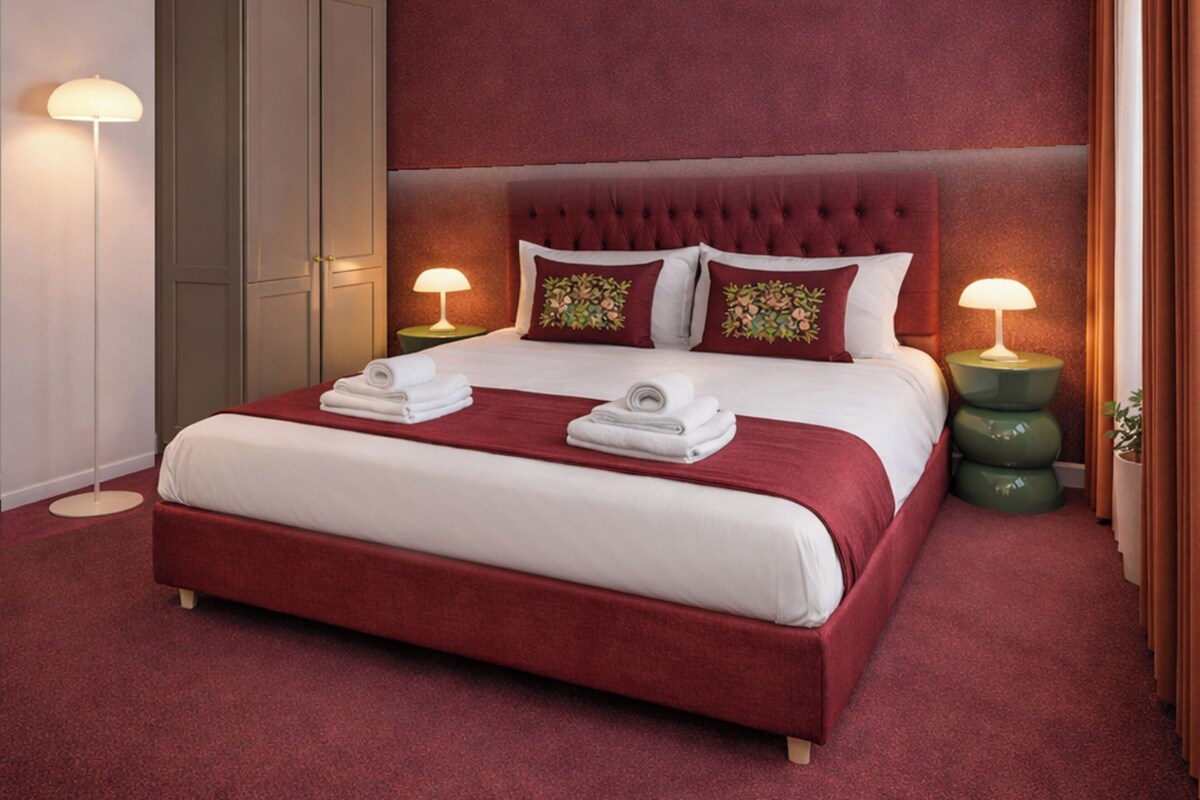 A hotel room with a large bed, two bedside tables with lamps, a wardrobe, and a red carpeted floor.