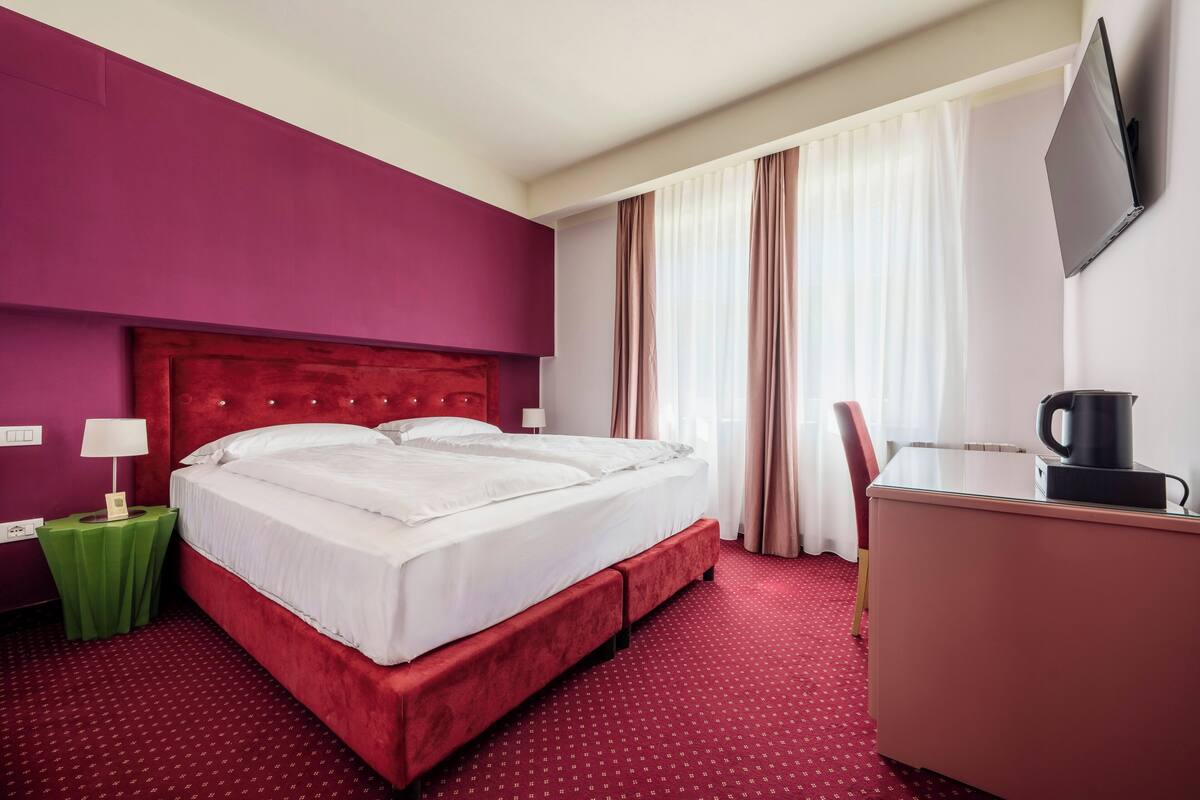 Ein Hotelzimmer mit einem roten Bett, einem grünen Nachttisch, einem Schreibtisch mit Kaffeemaschine und einem an der Wand montierten Fernseher.
