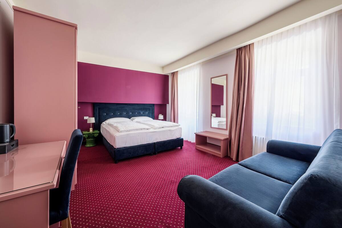 Ein Hotelzimmer mit einem Bett, einem Schreibtisch, einem Sessel und einer Couch, sowie einem großen Fenster mit Vorhängen.